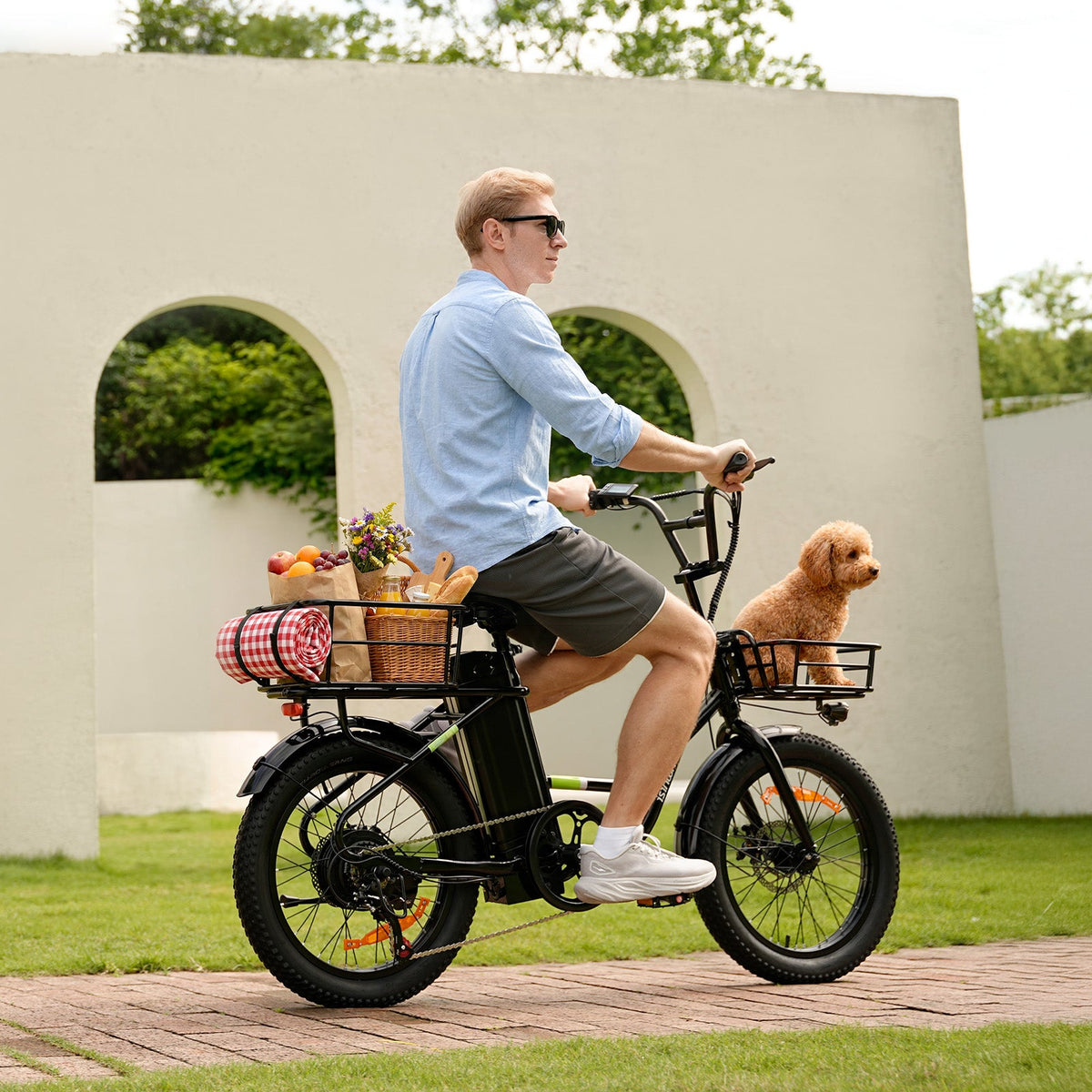 Rider on the isinwheel U7 in a park beside a white arched garden wall, with a picnic basket, fruit, and bread in the front basket and a small poodle in the rear basket
