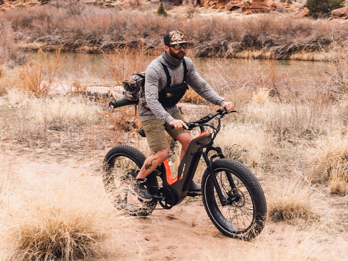 Rider in fishing gear on the Heybike Hero on a sandy dirt trail beside a river in a desert canyon, showing real-world off-pavement use