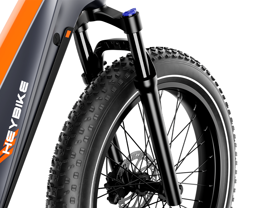 Detail view of the Heybike Hero front fork suspension stanchions, the front hydraulic disc brake caliper and rotor, and the front fat tire knobby tread