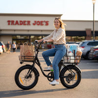 Rider seated on a black isinwheel U7 in front of a Trader Joe's storefront, with a Trader Joe's grocery bag in the front basket and milk, paper towels, and bread in the rear basket