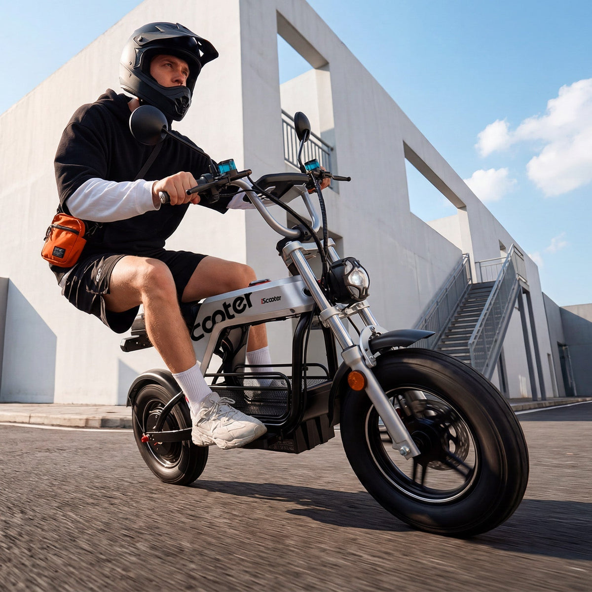 Rider on the iScooter DX5Pro outside a modern building with mirrors, headlight, padded seat, and broad deck visible.