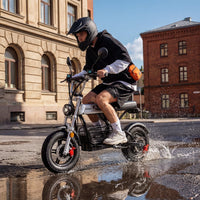 Helmeted rider taking the iScooter DX5Pro through a shallow puddle on city pavement, showing the seated stance and fat tires.