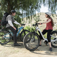 Two riders stopped with teal Dremax commuter e-bikes during a casual weekend ride.