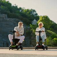 Two adults riding ScootNGo Lite mobility scooters on a paved waterfront path