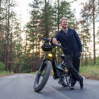 Rider standing beside a headlight-lit Nomad Pro in a forest at dusk.
