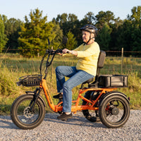 Older rider seated on the orange Meet One Breeze Pro 2.0 electric trike on a gravel path with front and rear baskets mounted.