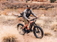 Rider in fishing gear on the Heybike Hero on a sandy dirt trail beside a river in a desert canyon, showing real-world off-pavement use