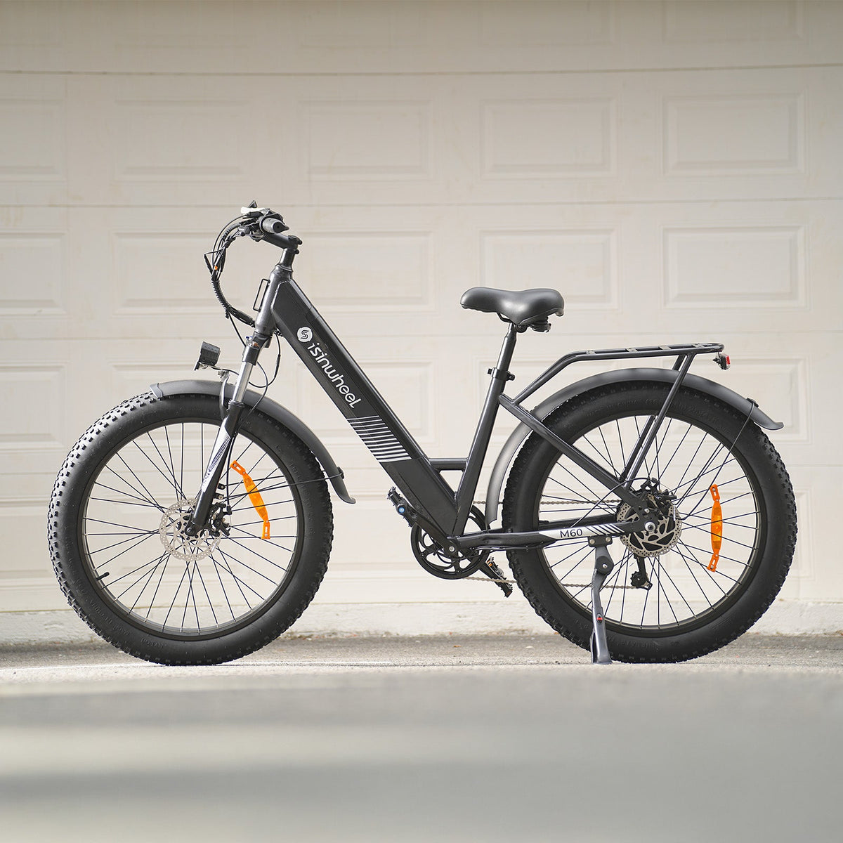 Iron Gray isinwheel M60 photographed parked in front of a residential garage door, showing the step-through frame, rear rack, fenders, padded saddle, and 26-inch fat tires
