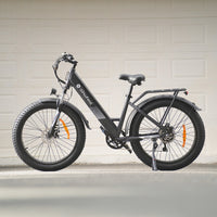 Iron Gray isinwheel M60 photographed parked in front of a residential garage door, showing the step-through frame, rear rack, fenders, padded saddle, and 26-inch fat tires