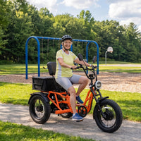 Orange Meet One Tour 2.0 semi-recumbent electric trike in side profile studio view, showing the recumbent saddle, the rear cargo basket with the backrest installed, the differential rear axle, and the 20 by 4 inch fat tires