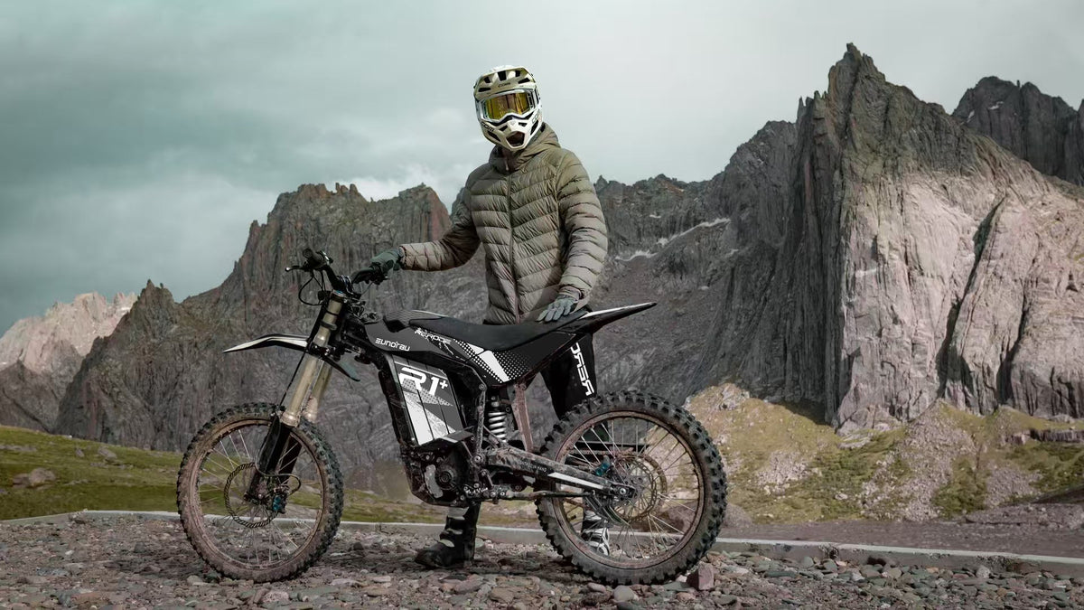 Helmeted rider beside a mud-splashed black R1+ electric dirt bike in rocky terrain