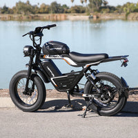 Black HappyRun G100 Pro Tank moto-styled electric bike on a paved promenade beside a lake, showing the long bench saddle, motorcycle-style handlebar, round caged LED headlight, dual rear shocks, and 20 by 4 inch road tires
